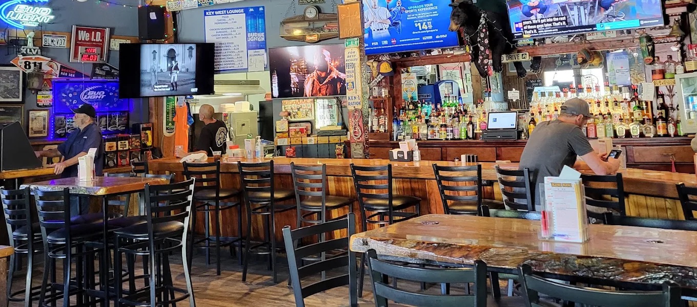 Inside Key West Lounge in North Adams, a wooden bar lined with stools, license plates on the walls, and TVs showing live sports above a full back bar